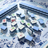 Blue-trimmed acrylic Mahjong tiles arranged in a wall formation with hand-illustrated character and flower tiles scattered on a chinoiserie table mat