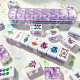 Purple marbled acrylic Mahjong tiles arranged in a wall with hand-illustrated wind, dragon and flower tiles laid out on a patterned mat