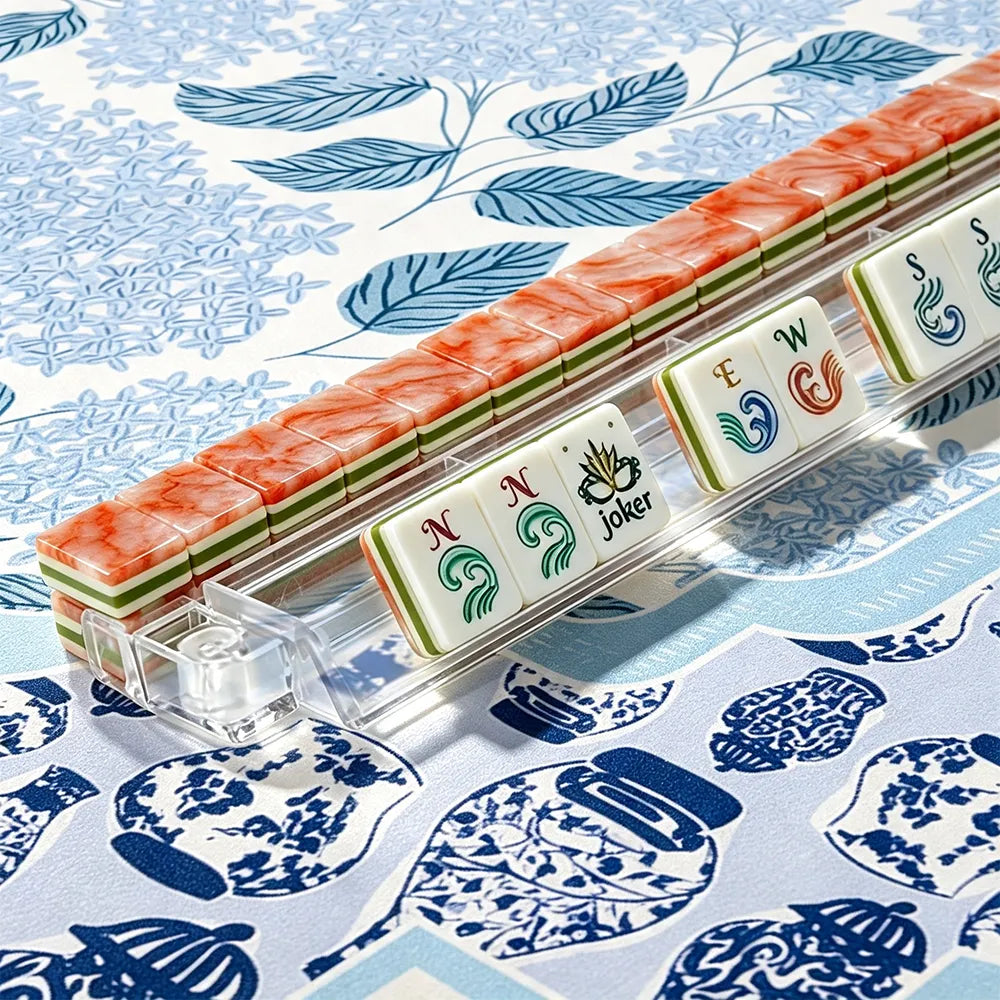 Coral marbled acrylic Mahjong tiles with hand-illustrated wind and joker tiles lined up in a clear tile rack on a blue chinoiserie mat