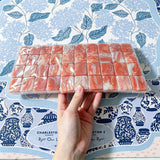 Hand holding a tray of coral and white marbled acrylic Mahjong tiles wrapped in plastic on a blue chinoiserie table mat