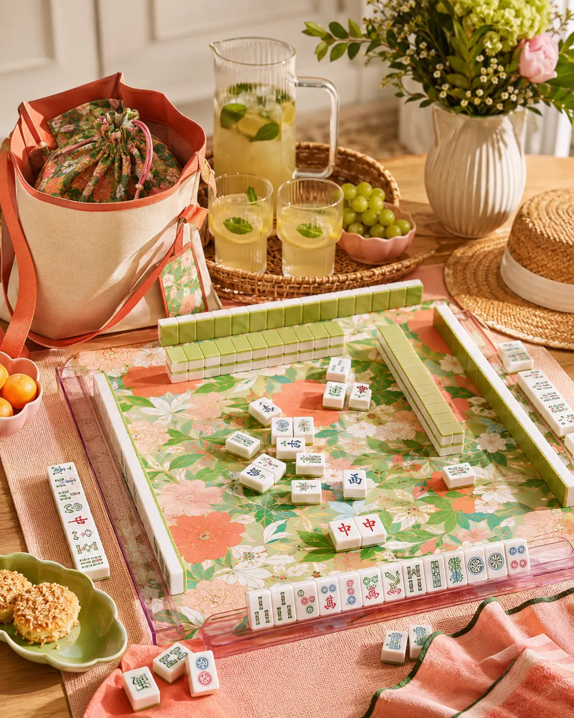 Cozy home mahjong tabletop scene with floral mat, green tile racks, lemonade, snacks, flowers, and a stylish tote bag.