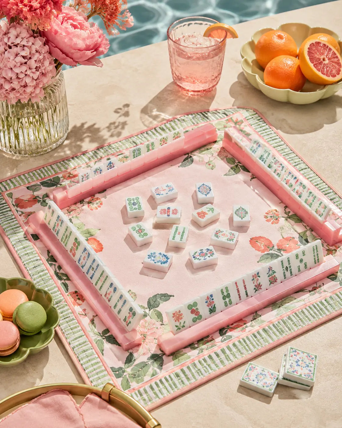 Pink floral mahjong set styled on a sunny poolside tabletop with pink racks, drinks, citrus fruit, flowers, and macarons.