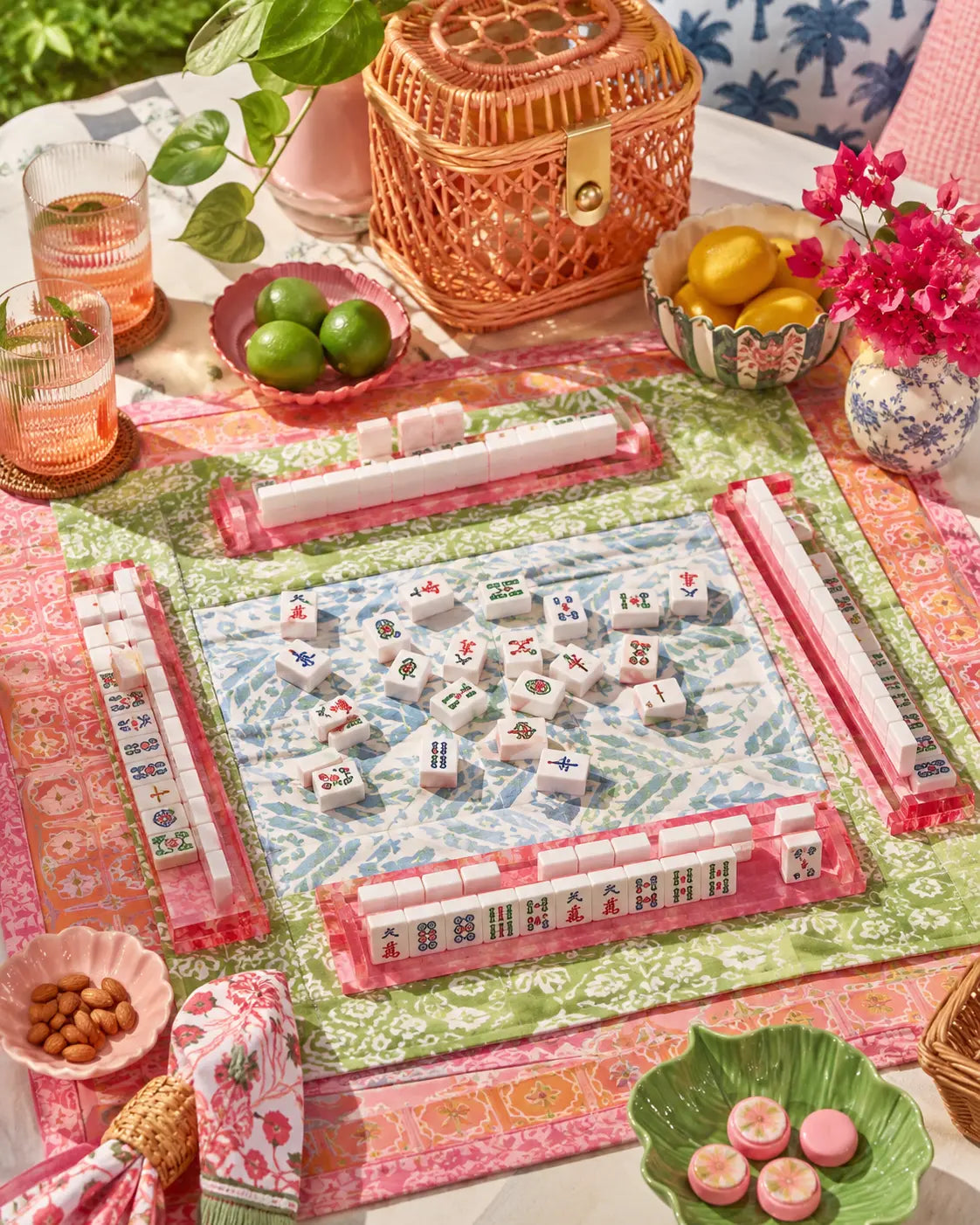 Colourful regional-style mahjong tabletop scene with patterned mats, pink racks, classic tiles, drinks, fruit, and floral decor.